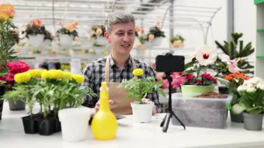 Bir adam serada video çekerken bir bitki gösteriyor. Çiçekler ve bahçe aletleri beyaz bir masada..