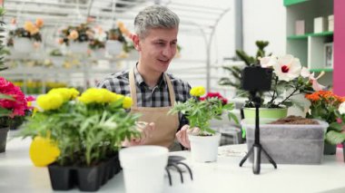 A man is recording a gardening video with plants and flowers in a greenhouse.
