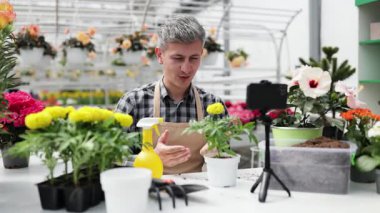 Seradaki bir adam, çiçeklerle ve bitkilerle çevrili telefonuyla bahçıvanlık dersi veriyor..