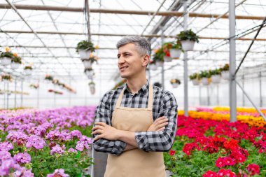 A smiling man in a greenhouse, surrounded by colorful flowers, wearing an apron and plaid shirt, looking content.