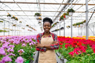 Gülümseyen Afrikalı bir kadın renkli çiçeklerle dolu bir serada duruyor..