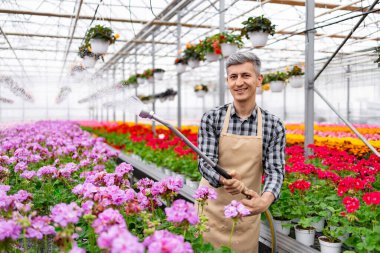 Gülümseyen bir adam, bahçeciliğe olan bağlılığını sergileyerek, bir serada sıra sıra renkli çiçekleri sular..