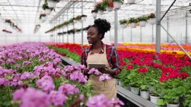 Bir tabletle bir serada gülümseyen Afrikalı kadın, canlı çiçekler ve bitkilerle çevrili, ve diğer saksı bitkileri.