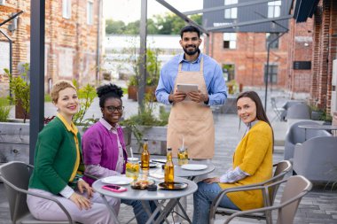 Bir grup arkadaş, açık hava kafesinde yemek yiyor. Dost canlısı bir garson servis ediyor..