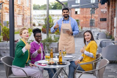 Bir grup mutlu arkadaş ve bir garson açık hava restoranında baş parmak gösteriyorlar..