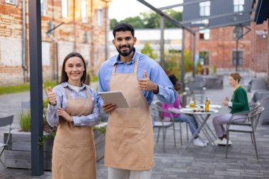 İki restoran çalışanı, bir erkek ve bir kadın, dışarıda poz veriyorlar. İkisi de gülümsüyor ve başparmak işareti yapıyor..