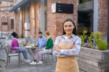 Bir restoran avlusunda kollarını kavuşturmuş gülümseyen garson, arka plandaki masada müşterilerle..