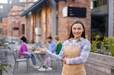 Önlük giymiş gülümseyen bir garson müşterileri açık hava restoranına davet eder. Diğer müşteriler arka plandaki masalarda otururlar..