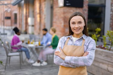 Önlük giymiş gülümseyen bir garson açık hava restoranının önünde poz veriyor, müşteriler arka plandaki masalarda oturuyor..