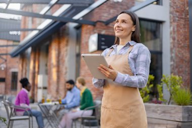 Bir garson elinde tablet, bakarken gülümsüyor, arkasında da restoran müşterileri var..