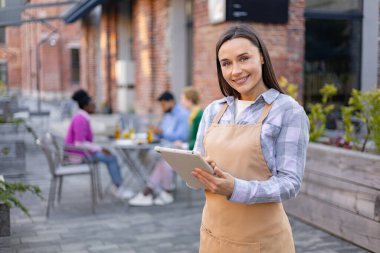 Restoranda gülümseyen bir garson elinde tabletle kameraya bakıyor. Lokantalar arka planda..