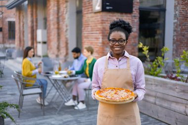 Gülümseyen Afrikalı garson elinde pizza, açık hava restoranında müşterilere servis yapıyor..