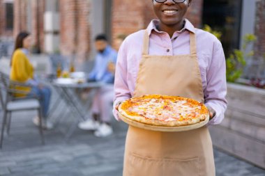 Dışarıdaki verandada pizza servisi yapan gülümseyen garson ve arka planda diğer müşteriler yemeklerini yiyor..