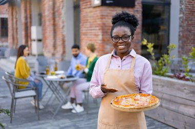 Gülümseyen bir garson dışarıda pizza tutuyor, müşterileri davet ediyor. Lokantalar bulanık arka planda masalarda otururlar.