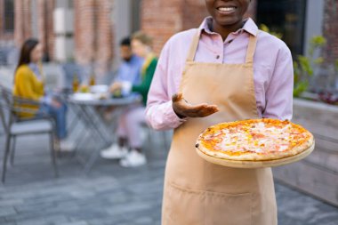 Gülümseyen bir garson, ahşap bir tahtada lezzetli bir pizza sunar. Müşterilere servis edilmeye hazırdır. Açık hava yemek deneyiminin tadını çıkarırlar..