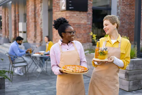 İki garson kız gülümsüyor, arka planda müşterileri olan bir restoranda pizza ve içki taşıyorlar..