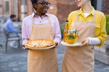 İki garson kız bir restoranda gülümsüyor ve ellerinde yiyecek ve içecekler var..