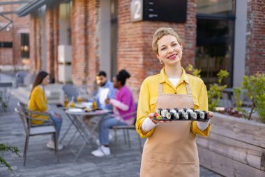 Gülümseyen bir garson verandada suşi sarmaları sunar. Müşteriler arka planda yemek yiyor..