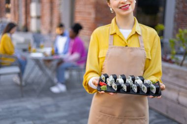 Gülümseyen bir garson dışarıda bir tabak suşi tutuyor. Lokantalar arka planda bulanık, yemeklerini yiyorlar..