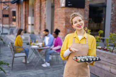 Gülümseyen garson, arka planda müşterileri olan açık hava restoranında bir tabak suşi sunar..