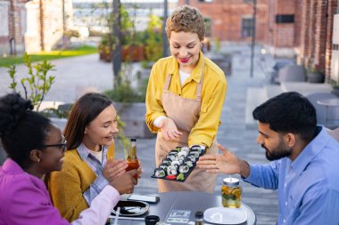 Bir garson dışarıda bir restoranda yemek yiyen bir grup arkadaşa suşi servis ediyor. Mutlular..