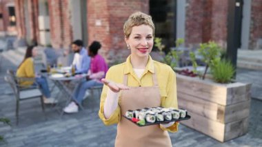 Dışarıdaki bir restoranda bir tabak suşi sunar. Gururla gülümserler. Konuklar arka planda görülebilir.