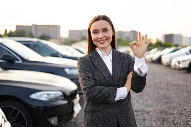Takım elbiseli bir kadın otoparkta dikilirken gülümsüyor, iyi bir jest yapıyor, başarılı bir alışveriş ya da satış öneriyor..