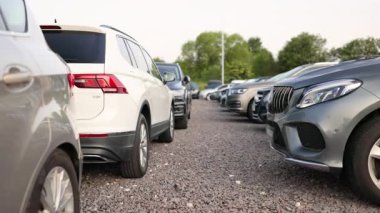 Dışarıda, çakıllı bir yüzeyde, satılığa hazır bir sıra farklı model araba. Araçlar iyi aydınlatılmış bir açık hava ortamında mevzilenmiş..