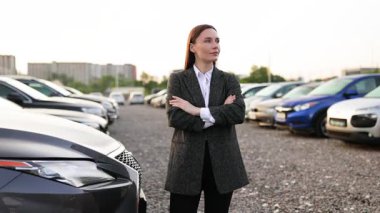 Gülümseyen bir iş kadını araba galerisinin önünde duruyor, kolları bağlı, araba satışlarında müşterilere yardım etmeye hazır..