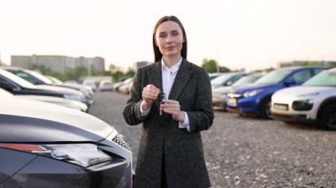 Araba anahtarlarını tutan, elinde tabletle gülümseyen takım elbiseli bir kadın bir araba galerisinde. Arabalar arka tarafa park edilmiş..