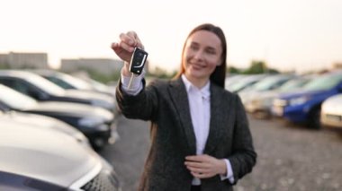 Gülümseyen bir kadın araba parkının önünde araba anahtarlarını tutuyor, muhtemelen bir araç aldıktan sonra..