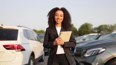 Afro-Amerikalı bir kadın, elinde tablet olan bir araba satıcısı, bir araba galerisinde başparmağıyla kameraya gülümsüyor..
