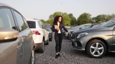 Takım elbiseli Afro-Amerikalı bir kadın elinde tablet, bir galeride arabayı sunuyor, potansiyel bir alıcıya özelliklerini gösteriyor..