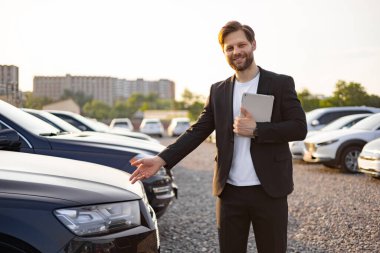 Takım elbiseli gülümseyen bir araba satıcısı elinde tabletle yeni bir arabaya doğru hareket eder. Arka planda gün batımının altında diğer araçlar görünür..