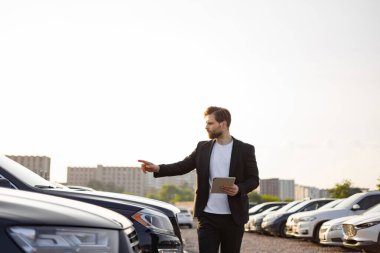 Takım elbiseli bir işadamı park yerindeki bir arabayı işaret ediyor. Elinde tablet var ve araçları inceliyor..