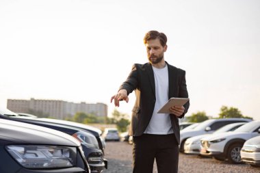 Takım elbiseli bir araba satıcısı güneşli bir günde bir otoparkta tablet tutarken bir aracı işaret ediyor..