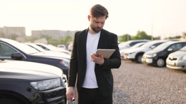 Gülümseyen bir araba satıcısının elinde anahtarlar, bir otoparkta duruyor, çeşitli araçlarla çevrili, araba satın alma ya da sahiplik öneriyor..