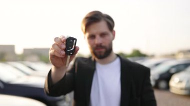 Bir işadamı kameraya gülümseyen bir araba anahtarı tutuyor. Arka planda arabaları olan bir araba galerisinde görünüyor..