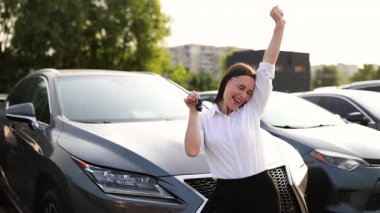 Neşeli bir kadın araba alımını kutluyor, heyecandan anahtarları tutuyor, bir araba galerisinde yeni aracın yanında duruyor..