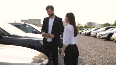 Bir araba satıcısı, güneşli bir günde bir kadın müşterisiyle arabaların arasında evrakları gözden geçirir..