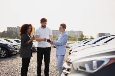 Bir araba satıcısı müşteriye alışverişte yardım ederken, diğer bir kadın da güneşli bir günde araba galerisinde izliyor..