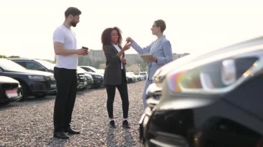 Bir kadın yeni araba alımını yakınlarda bir satıcı ve ortağıyla kutluyor. Arka planda çeşitli modellerin arabaları var..