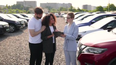 Bir araba satıcısı, çeşitli araçlarla çevrili bir araba galerisinde farklı bir çifte tabletle yardım eder..
