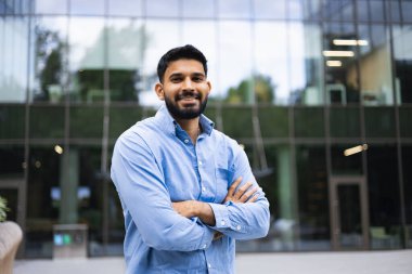 Gülümseyen Hintli bir adam, modern cam bir binanın önünde kollarını kavuşturarak güvenle duruyor..