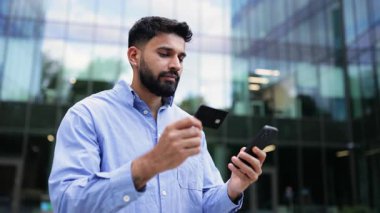 Elinde kredi kartı ve akıllı telefon tutan gülümseyen Hintli bir adam, muhtemelen modern bir binanın önünde online ödeme yapıyor..