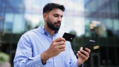 Hintli bir adam elinde bir kredi kartı ve akıllı bir telefonla dijital bankacılık ve finansal işlemleri temsil eden modern bir binanın önünde gülümsüyor..