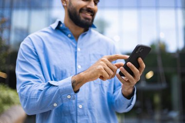 Sakallı, gülümseyen bir adam, arka planı bulanık, modern teknolojiyi sergileyen bir akıllı telefon kullanıyor..