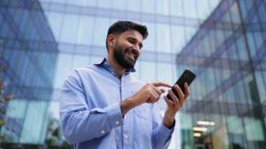 Mavi gömlekli Hintli bir adam modern bir binanın önünde telefonunu kontrol ediyor..