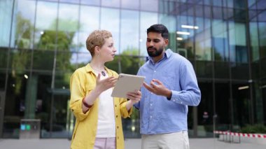 İki farklı iş arkadaşı modern bir ofis binasının önünde duruyor, gülümsüyor ve kameraya bakıyor, biri tablet tutuyor..