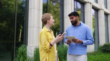Bir kadın telefonuyla selfie çekerken, bir erkek barış işareti yapıyor ve açık havada tablet tutuyor. Gülümsüyorlar..